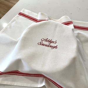 May include: White linen bread cloth with red stripes and the embroidered text "Ashlyn's Sourdough". The cloth is folded over a round object, likely a loaf of bread, and is placed on a light-colored surface.