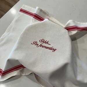 Puede incluir: Pa&ntilde;o de cocina de lino blanco con rayas rojas y el texto bordado "Shhh... I'm fermenting". El pa&ntilde;o est&aacute; doblado sobre otro pa&ntilde;o con rayas rojas, lo que sugiere un tema de cocina o reposter&iacute;a.