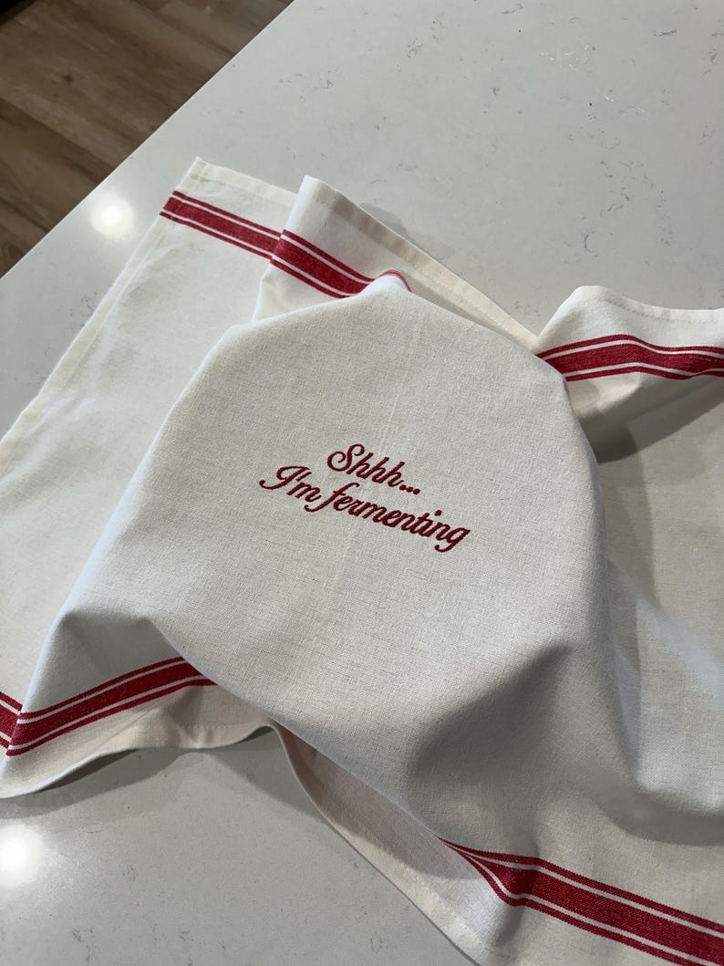 Puede incluir: Pa&ntilde;o de cocina de lino blanco con rayas rojas y el texto bordado "Shhh... I'm fermenting". El pa&ntilde;o est&aacute; doblado sobre otro pa&ntilde;o con rayas rojas, lo que sugiere un tema de cocina o reposter&iacute;a.
