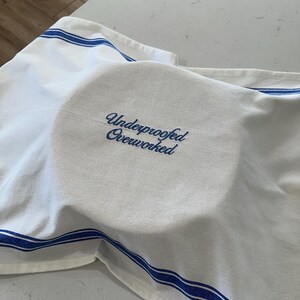 Puede incluir: Pa&ntilde;o de cocina blanco con rayas azules, que cubre un objeto redondo. El pa&ntilde;o tiene el texto bordado "Underproofed Overworked" en azul. El pa&ntilde;o est&aacute; doblado sobre el objeto.