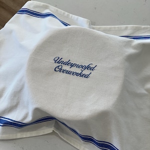 Puede incluir: Pa&ntilde;o de cocina blanco con rayas azules y el texto bordado "Underproofed Overworked" en azul. El pa&ntilde;o est&aacute; cubriendo un objeto redondo, posiblemente masa o un plato.