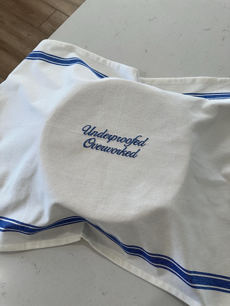 Puede incluir: Pa&ntilde;o de cocina blanco con rayas azules y el texto bordado "Underproofed Overworked" en azul. El pa&ntilde;o est&aacute; cubriendo un objeto redondo, posiblemente masa o un plato.
