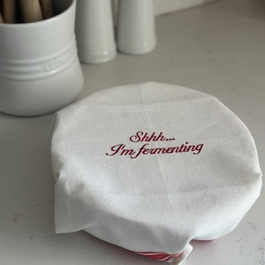 May include: A white fabric bowl cover with red embroidered text that reads "Shhh... I'm fermenting". The circular cloth features a red striped pattern visible at the bottom edge and is designed to fit snugly over the top of a container.