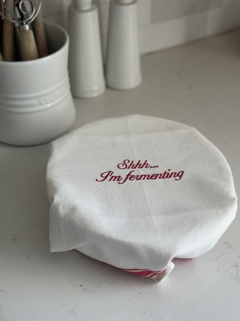 May include: A white fabric bowl cover with red embroidered text that reads "Shhh... I'm fermenting". The circular cloth features a red striped pattern visible at the bottom edge and is designed to fit snugly over the top of a container.