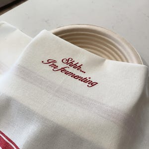 May include: A cream-colored cotton kitchen towel with red stripes along the bottom edge, featuring the embroidered red text "Shhh... I'm fermenting" in a cursive script, draped over a round, light-colored, ribbed cane proofing basket.