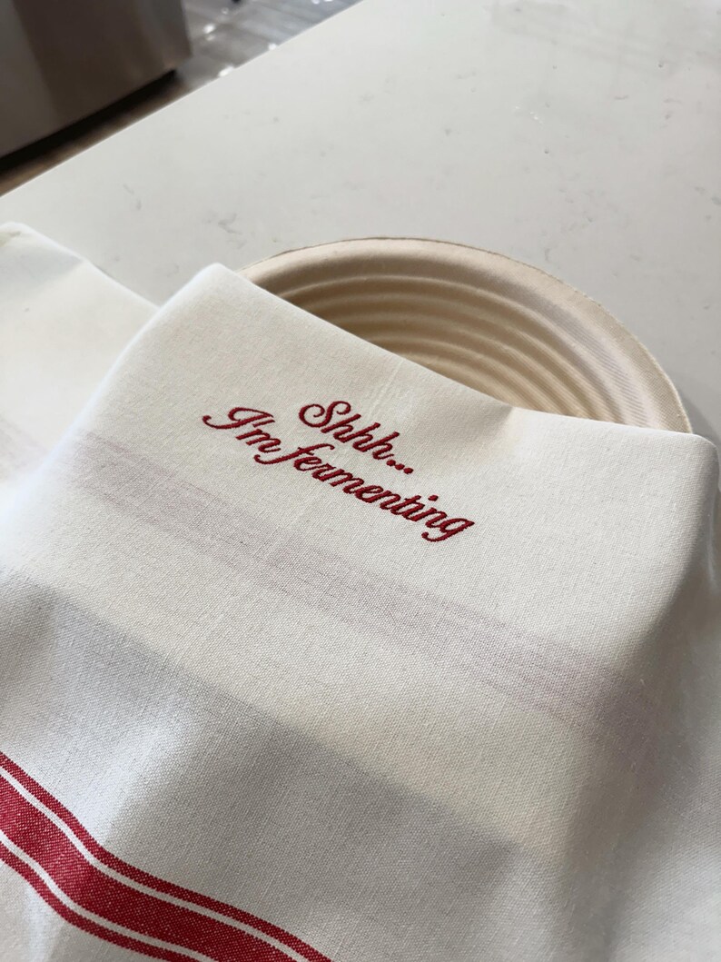May include: A cream-colored cotton kitchen towel with red stripes along the bottom edge, featuring the embroidered red text "Shhh... I'm fermenting" in a cursive script, draped over a round, light-colored, ribbed cane proofing basket.