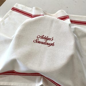 Puede incluir: Pa&ntilde;o de pan de lino blanco con rayas rojas y el texto bordado "Ashlyn's Sourdough". El pa&ntilde;o est&aacute; drapeado sobre un pan, lo que sugiere un tema de cocina o panader&iacute;a.