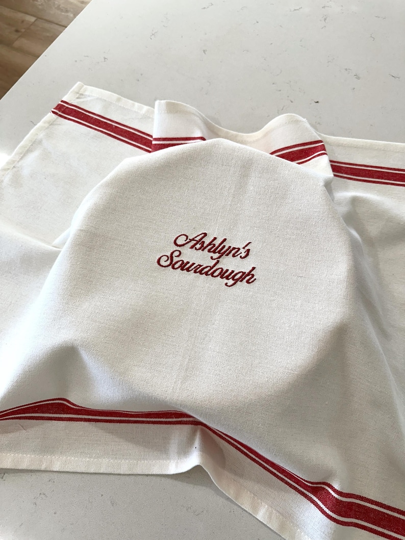 Puede incluir: Pa&ntilde;o de pan de lino blanco con rayas rojas y el texto bordado "Ashlyn's Sourdough". El pa&ntilde;o est&aacute; drapeado sobre un pan, lo que sugiere un tema de cocina o panader&iacute;a.