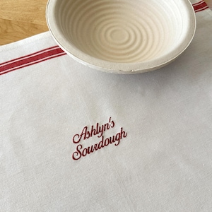 Puede incluir: Una cesta de fermentaci&oacute;n redonda beige descansa sobre un pa&ntilde;o de lino blanco con rayas rojas y el texto bordado "Ashlyn's Sourdough". La cesta tiene ranuras circulares conc&eacute;ntricas.
