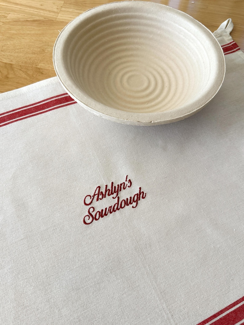 Puede incluir: Una cesta de fermentaci&oacute;n redonda beige descansa sobre un pa&ntilde;o de lino blanco con rayas rojas y el texto bordado "Ashlyn's Sourdough". La cesta tiene ranuras circulares conc&eacute;ntricas.