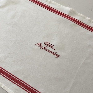 May include: A rectangular off-white kitchen towel featuring two horizontal red stripes near the top and bottom edges. The center of the towel is embroidered with the red text "Shhh... I'm fermenting" in a cursive script font style.