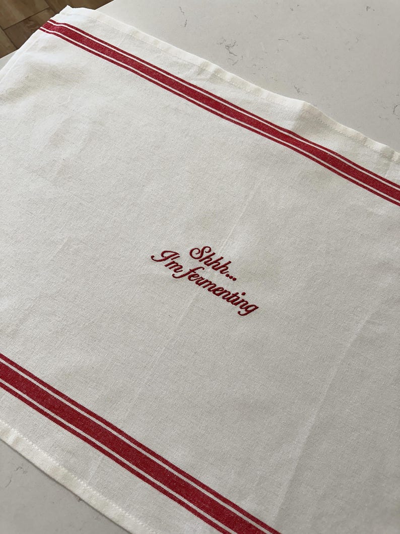 May include: A rectangular off-white kitchen towel featuring two horizontal red stripes near the top and bottom edges. The center of the towel is embroidered with the red text "Shhh... I'm fermenting" in a cursive script font style.