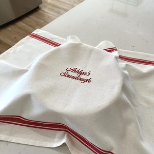 Puede incluir: Toalla de cocina blanca con rayas rojas y el texto bordado "Ashlyn's Sourdough". La toalla est&aacute; colocada sobre un cuenco en una encimera blanca. La toalla est&aacute; hecha de un material suave y absorbente.
