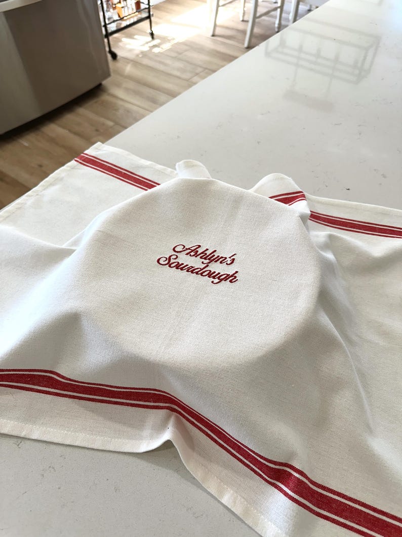 Puede incluir: Toalla de cocina blanca con rayas rojas y el texto bordado "Ashlyn's Sourdough". La toalla est&aacute; colocada sobre un cuenco en una encimera blanca. La toalla est&aacute; hecha de un material suave y absorbente.