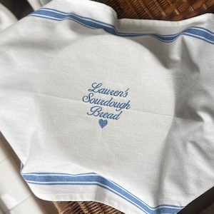 May include: A white kitchen towel featuring blue striped borders and blue embroidery that reads "Lauren's Sourdough Bread" above a small heart icon. The fabric is draped, showing the text clearly centered on the white cotton material.