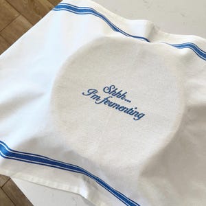 May include: A white kitchen towel with blue striped borders and the embroidered blue text Shhh... I'm fermenting. The fabric is draped over a sourdough banneton basket, creating soft folds and shadows across the surface of the textile material.