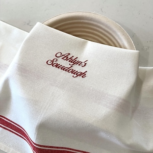 Puede incluir: Pa&ntilde;o de cocina de lino blanco con rayas rojas y texto bordado "Ashlyn's Sourdough". El pa&ntilde;o est&aacute; doblado sobre una cesta de pan redonda beige, lo que sugiere un entorno de cocina o panader&iacute;a. El dise&ntilde;o es simple y cl&aacute;sico.