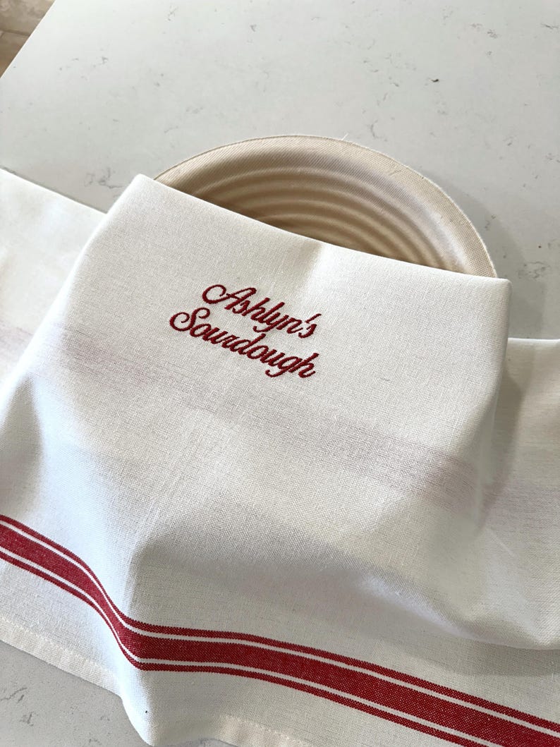 Puede incluir: Pa&ntilde;o de cocina de lino blanco con rayas rojas y texto bordado "Ashlyn's Sourdough". El pa&ntilde;o est&aacute; doblado sobre una cesta de pan redonda beige, lo que sugiere un entorno de cocina o panader&iacute;a. El dise&ntilde;o es simple y cl&aacute;sico.