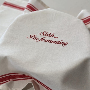 May include: An off-white kitchen towel featuring red striped borders and the embroidered red text "Shhhh... I'm fermenting" centered on the fabric. The towel is draped over an object, displaying its woven texture and the decorative red lines.