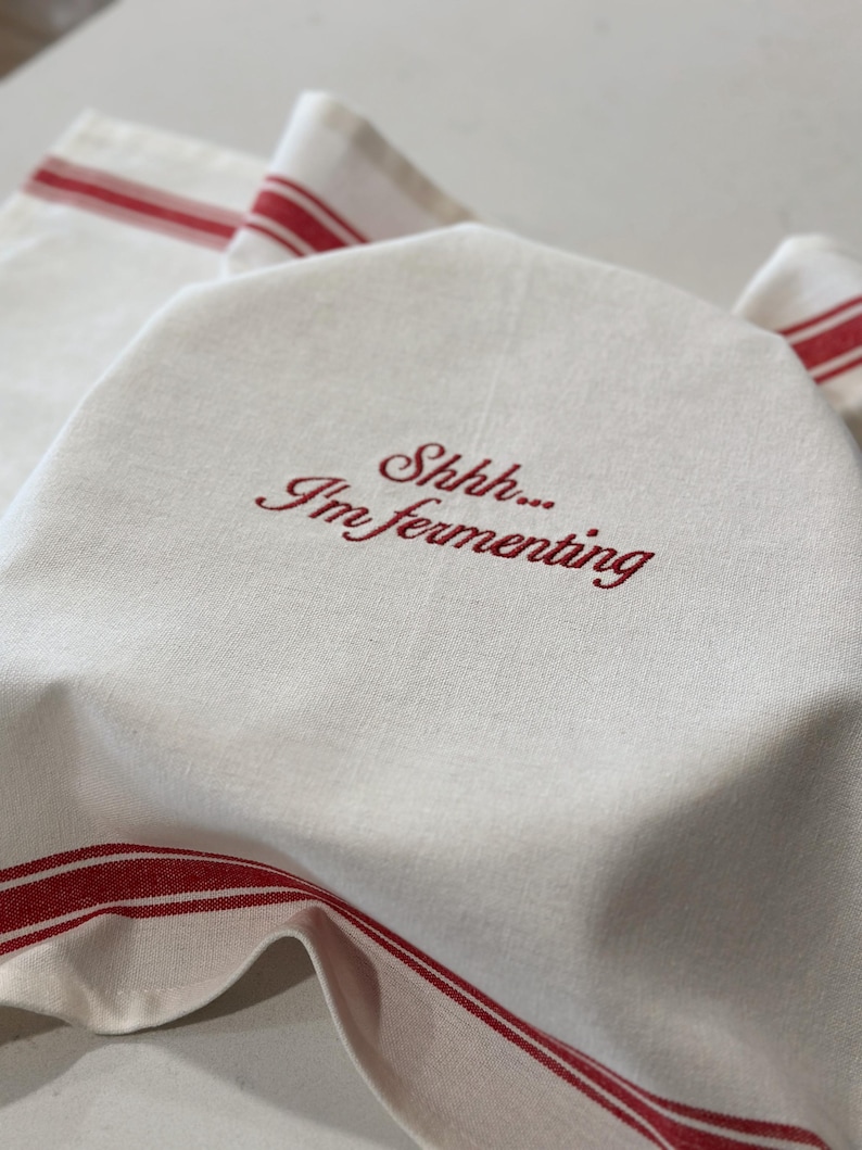 May include: An off-white kitchen towel featuring red striped borders and the embroidered red text "Shhhh... I'm fermenting" centered on the fabric. The towel is draped over an object, displaying its woven texture and the decorative red lines.