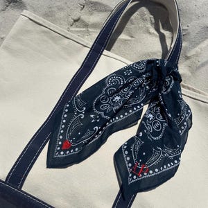 May include: A cream-colored canvas tote bag with dark navy blue straps and base, featuring a dark navy blue bandana tied to the handle. The bandana has a white paisley pattern, a small red heart embroidery, and red embroidered initials.