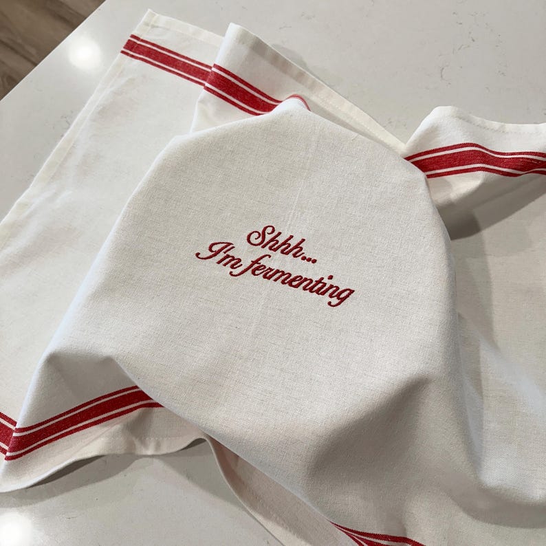 May include: A white kitchen towel with red striped borders and the embroidered red text Shhh... I'm fermenting. The fabric is draped over an object, creating soft folds and shadows across the surface of the textile material.