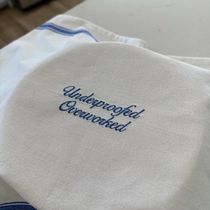 Puede incluir: Pa&ntilde;o de cocina blanco con rayas azules y el texto bordado "Underproofed Overworked" en letra cursiva. El pa&ntilde;o est&aacute; doblado, con el texto en un lugar destacado.