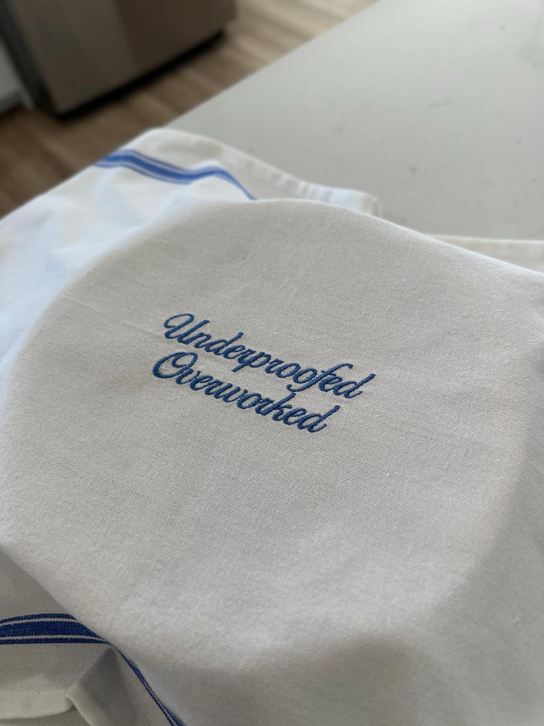 Puede incluir: Pa&ntilde;o de cocina blanco con rayas azules y el texto bordado "Underproofed Overworked" en letra cursiva. El pa&ntilde;o est&aacute; doblado, con el texto en un lugar destacado.