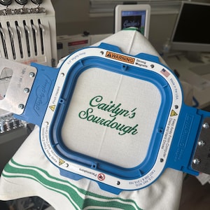 Puede incluir: Un marco de bordado azul Mighty Hoop que sostiene una tela blanca con el texto verde "Caitlyn's Sourdough". El marco tiene una etiqueta de advertencia y est&aacute; hecho en los EE. UU. La tela tiene rayas verdes.
