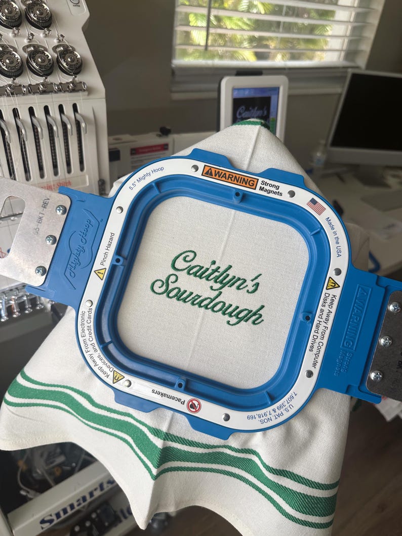 Puede incluir: Un marco de bordado azul Mighty Hoop que sostiene una tela blanca con el texto verde "Caitlyn's Sourdough". El marco tiene una etiqueta de advertencia y est&aacute; hecho en los EE. UU. La tela tiene rayas verdes.