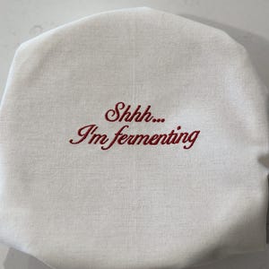 May include: A round, off-white fabric cover featuring the embroidered red text "Shhh... I'm fermenting" in a cursive script. The cover has a gathered edge, designed to fit over the top of a jar or container to protect its contents during the process.