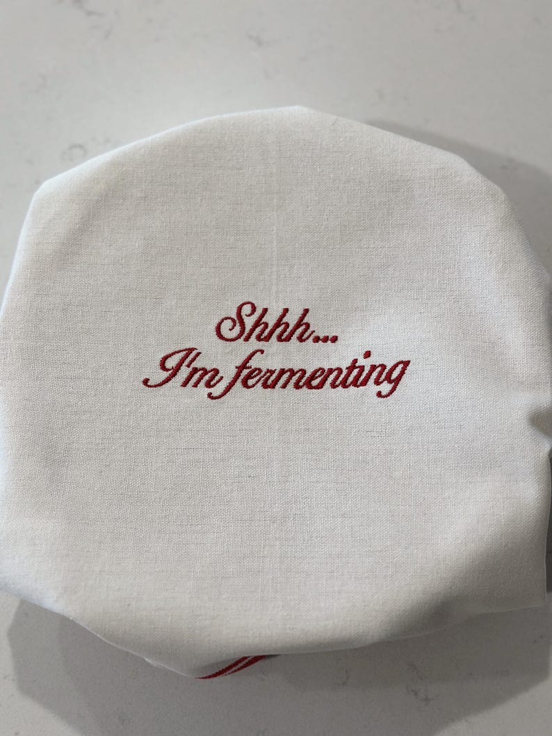 May include: A round, off-white fabric cover featuring the embroidered red text "Shhh... I'm fermenting" in a cursive script. The cover has a gathered edge, designed to fit over the top of a jar or container to protect its contents during the process.