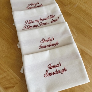 Puede incluir: Cuatro pa&ntilde;os de cocina blancos con bordados burdeos. Los pa&ntilde;os tienen los textos: "Anna's Sourdough", "I like my bread like I like my Jesus... risen!", "Shelby's Sourdough" y "Jenna's Sourdough".