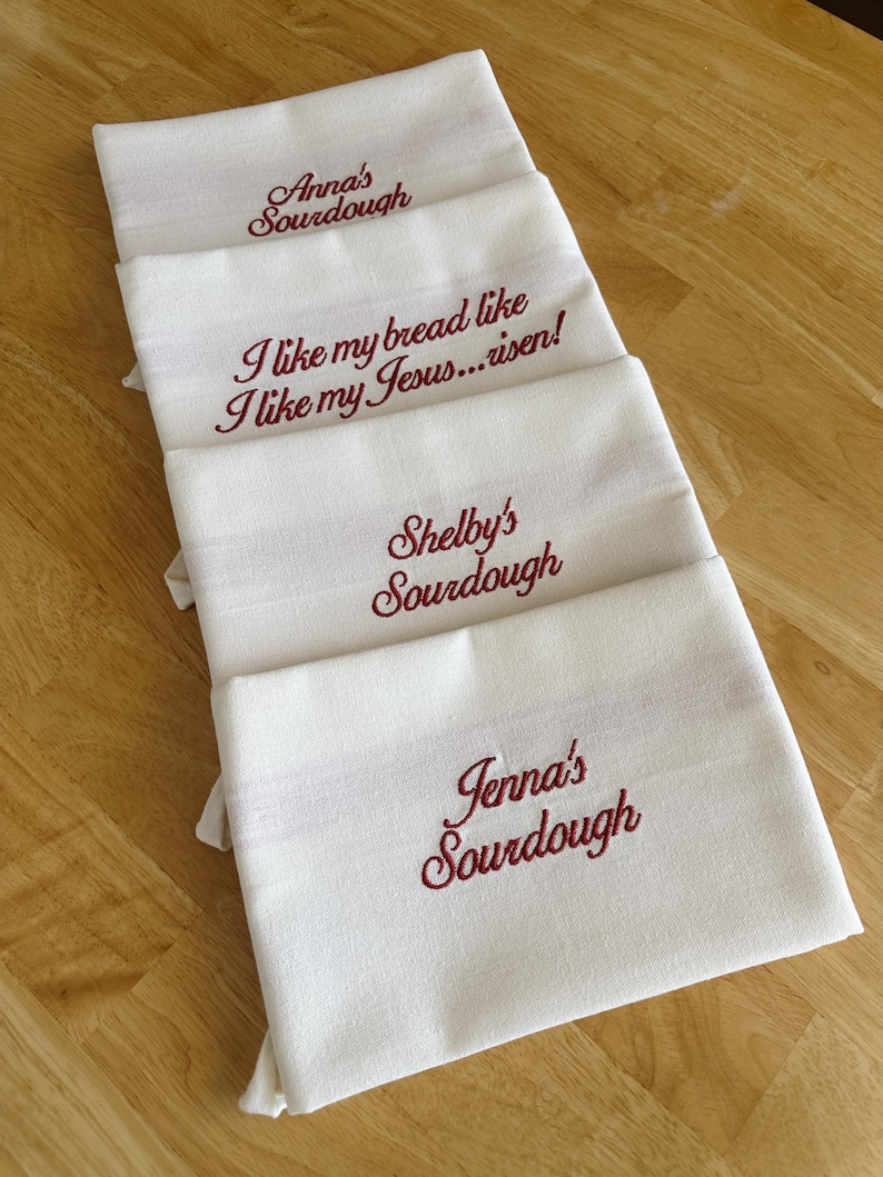 Puede incluir: Cuatro pa&ntilde;os de cocina blancos con bordados burdeos. Los pa&ntilde;os tienen los textos: "Anna's Sourdough", "I like my bread like I like my Jesus... risen!", "Shelby's Sourdough" y "Jenna's Sourdough".