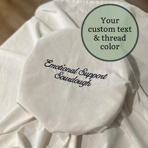 May include: Off-white, octagonal bread cover with the embroidered text "Emotional Support Sourdough" in dark blue script. A circular graphic in the upper right corner reads "Your custom text & thread color". The cover is on a white cloth.