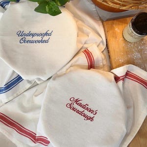May include: Two off-white cotton bowl covers with elasticated edges. One cover features blue embroidered text reading Underproofed Overworked, and the other features red embroidered text reading Madison's Sourdough. Both have striped borders.