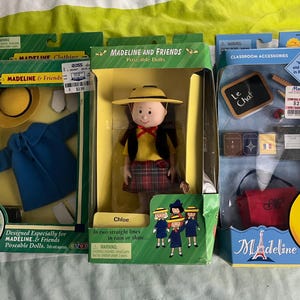 May include: Three Madeline doll sets, one with a red and black plaid dress, one with a blue coat, and one with classroom accessories. The sets are packaged in boxes with the Madeline logo and the words "Poseable Dolls" and "Classroom Accessories".