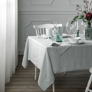 May include: A white table with a light gray linen tablecloth, set with white plates, glasses, and a white pitcher. The table is decorated with a green glass bottle and a white pitcher. The table is set for a formal dinner.