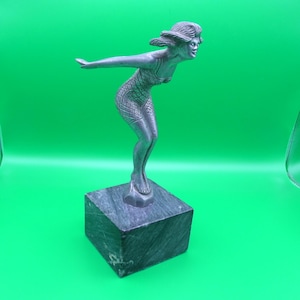 May include: A silver-toned sculpture of a woman in a diving pose, mounted on a square, dark gray marble base. The woman is wearing a patterned swimsuit and has her arms outstretched. The sculpture has a vintage, art deco style.
