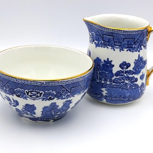 May include: A blue and white ceramic creamer and sugar bowl set. The pieces feature a classic Willow pattern design with gold trim. The creamer has a handle and spout, while the bowl is round.