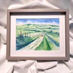 May include: Watercolour painting of a green countryside scene with a track and a fence. The painting is framed in a grey frame with a white mount. The sky is blue with white clouds. Two figures walk down the path.