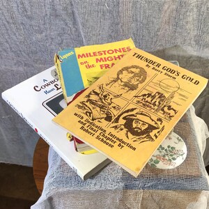 May include: A stack of three paperback books with yellow, white, and brown covers. The top book is titled "Thunder God's Gold" by Barry Storm. The middle book is titled "Milestones on the Mighty Frontier". The bottom book is titled "A Cowboy Never Lies".