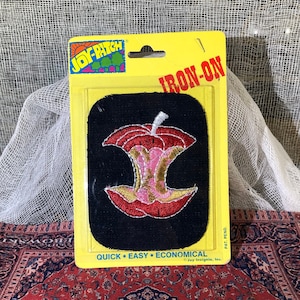 May include: A red and yellow iron-on patch of a bitten apple on a black background. The packaging says "Joy-Patch Iron-On" and "Quick Easy Economical".