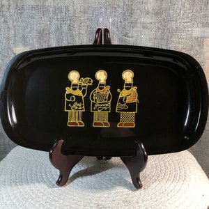 Puede incluir: Una bandeja rectangular negra con esquinas redondeadas. La bandeja presenta tres figuras de chef doradas con las palabras "Chef", "Gourmet" y "Cook" debajo. La bandeja se exhibe sobre un soporte de madera.