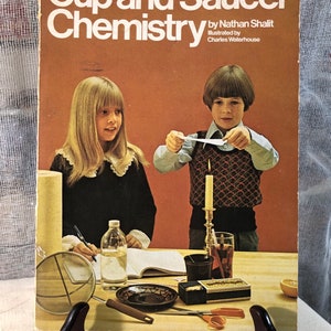 May include: A vintage book titled "Cup and Saucer Chemistry" by Nathan Shailit, illustrated by Charles Waterhouse. The cover features two children performing a science experiment with a candle and a piece of paper.