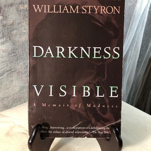 May include: A paperback book titled "Darkness Visible: A Memoir of Madness" by William Styron. The book cover features a dark brown background with the title in white and green text. The book is on a wooden book stand.