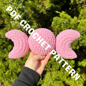 Puede incluir: Un patrón de ganchillo rosa con forma de triple luna, con el texto "PDF CROCHET PATTERN" en blanco. El ganchillo es de un material suave y texturizado. El fondo es un arbusto verde y frondoso.