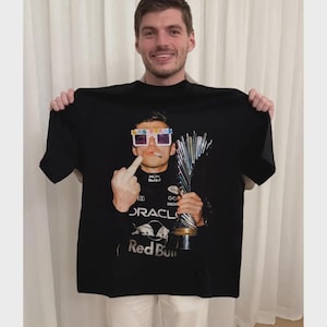 Puede incluir: Una camiseta negra de manga corta con un estampado gr&aacute;fico de una persona con gafas de sol de Las Vegas, sosteniendo un trofeo y haciendo un gesto con el dedo coraz&oacute;n. El estampado incluye el logo de Oracle Red Bull Racing.