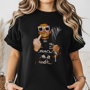Puede incluir: Una camiseta negra con un estampado gr&aacute;fico de un piloto de carreras que lleva gafas blancas de Las Vegas, sostiene un trofeo y hace un gesto con el dedo coraz&oacute;n. El piloto viste un traje de carreras negro Oracle Red Bull con logos.