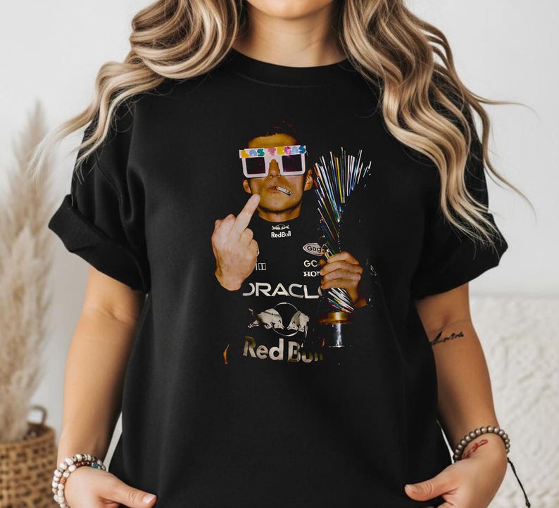 Puede incluir: Una camiseta negra con un estampado gr&aacute;fico de un piloto de carreras que lleva gafas blancas de Las Vegas, sostiene un trofeo y hace un gesto con el dedo coraz&oacute;n. El piloto viste un traje de carreras negro Oracle Red Bull con logos.